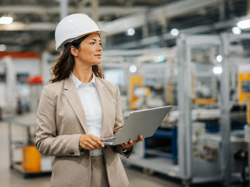 Vrouw met witte veiligheidshelm en laptop in een productiehal