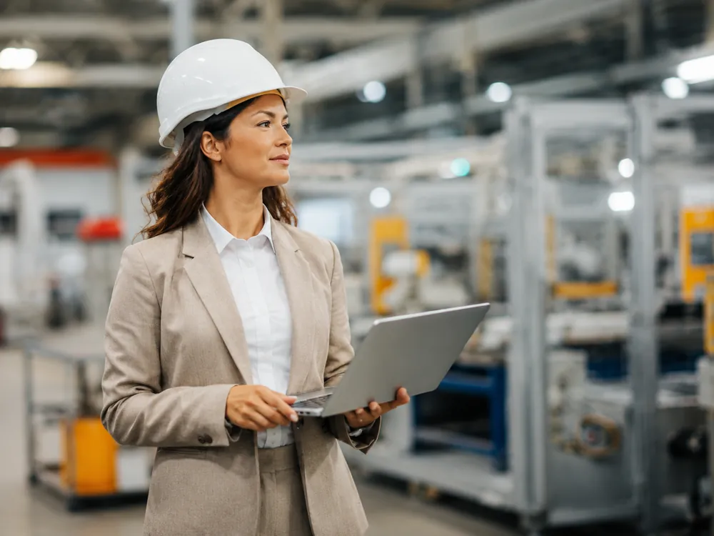 Vrouw met witte veiligheidshelm en laptop in een productiehal